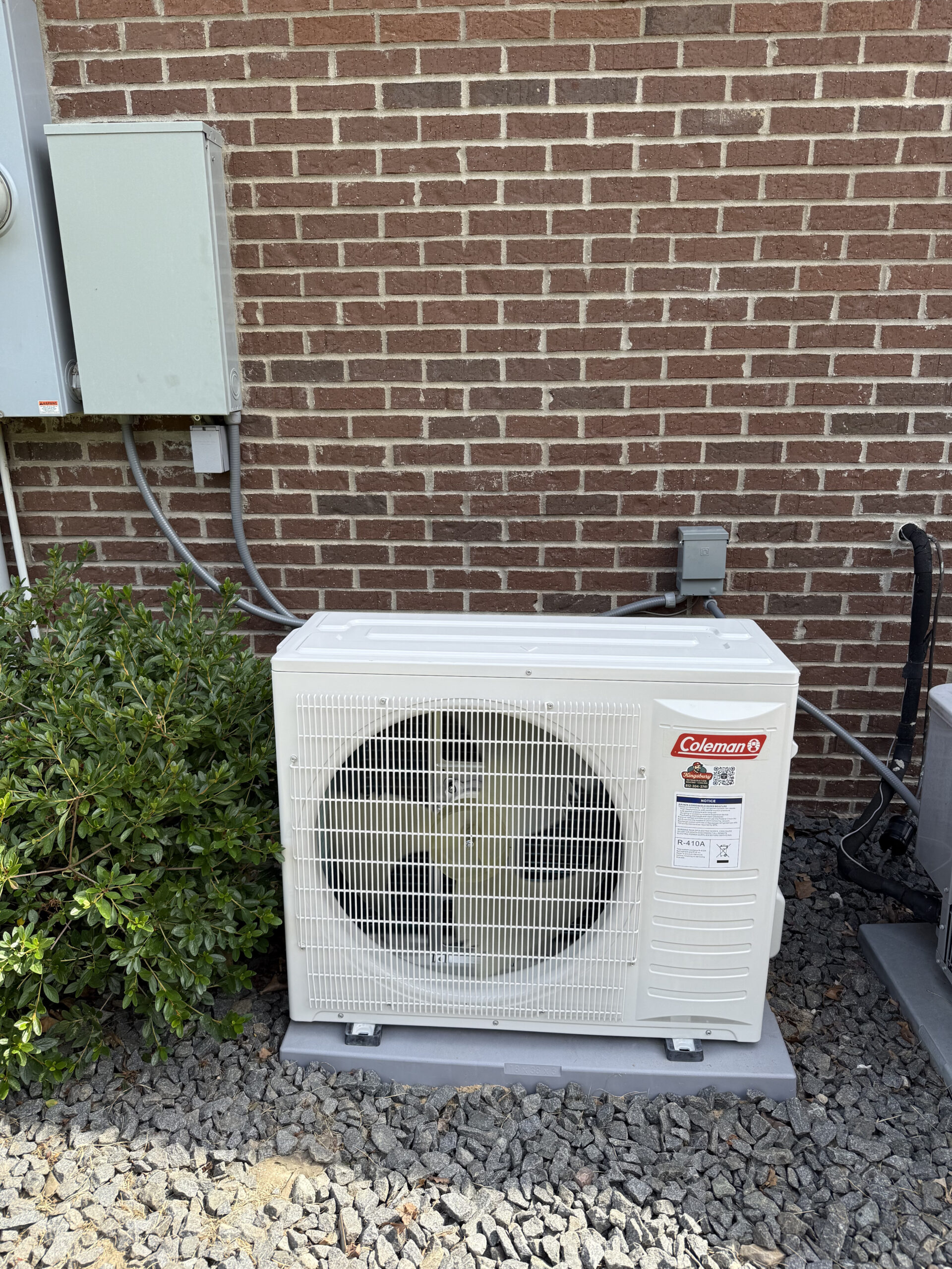 A white Coleman outdoor air conditioning unit sits on a concrete pad next to a brick wall, near an electrical box and some green bushes.