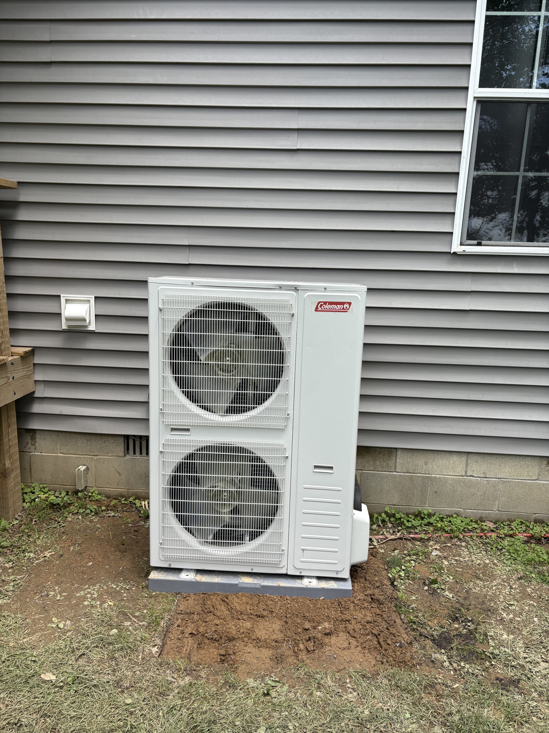 A white outdoor heat pump unit is installed on a concrete pad beside a house with gray siding and a window.