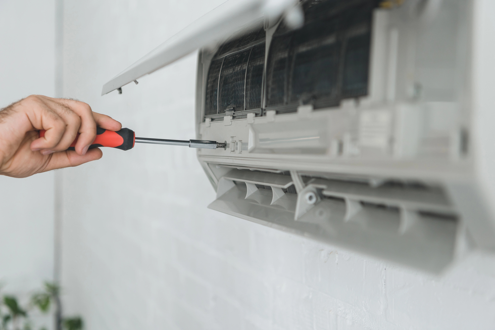 A person uses a screwdriver to repair or maintain an air conditioning unit mounted on a white wall.