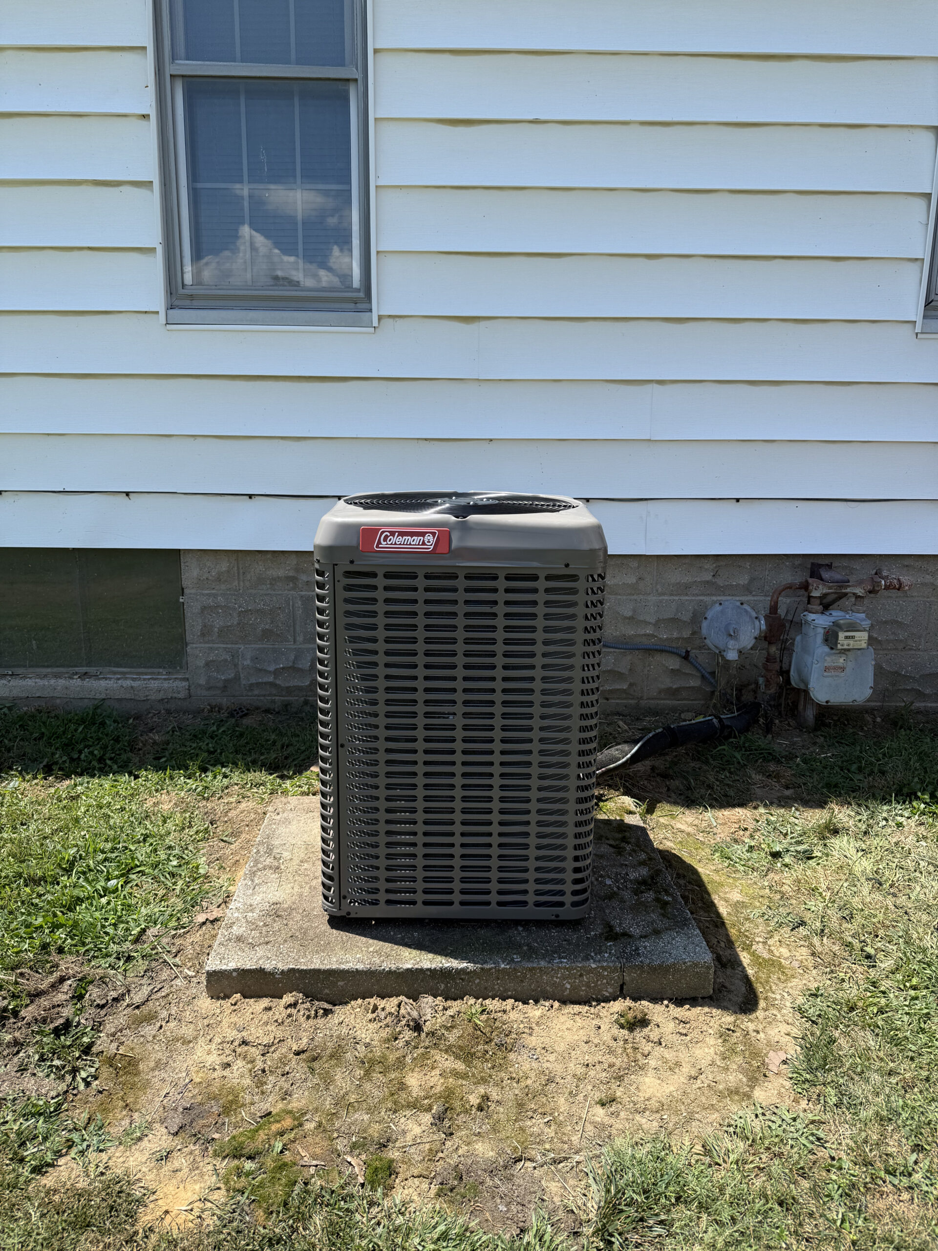 Coleman air conditioning unit sits on a concrete pad outside a white house with a window and siding visible above. Grass and exposed soil surround the unit.