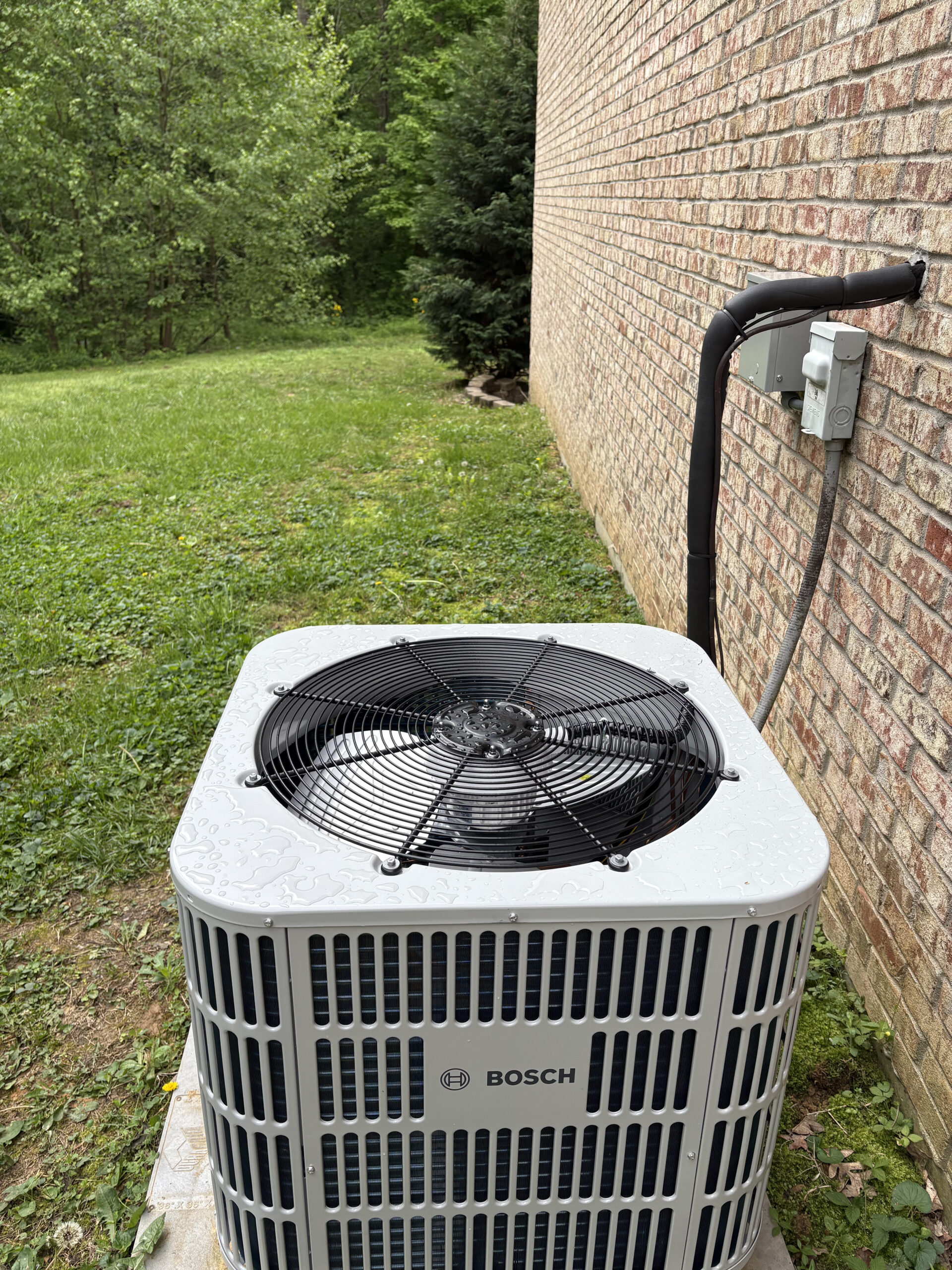A Bosch outdoor air conditioning unit is installed next to a brick house, with electrical connections running along the wall. A grassy yard and trees are in the background.