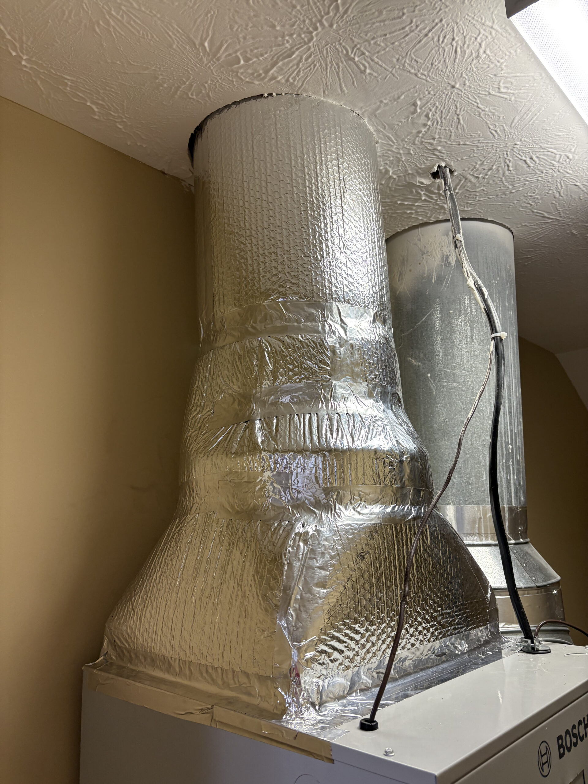 Insulated metal ductwork covered in reflective foil connects to a ceiling vent above a large appliance in a utility room.