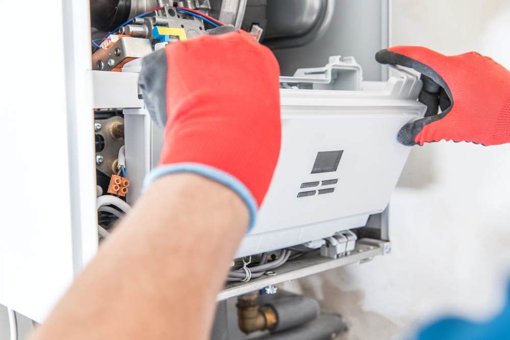 A person wearing red gloves is removing or installing the front panel of a gas boiler for maintenance or repair.